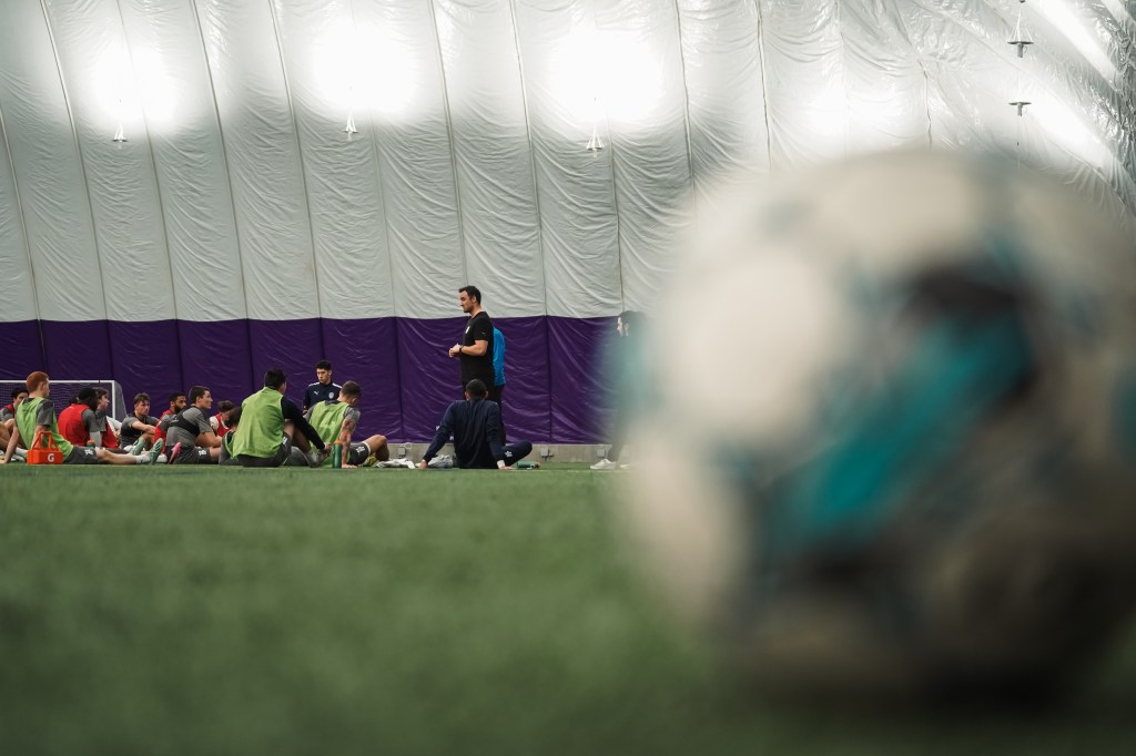 Head coach Eamon Zayed speaks to the team during a preseason training session. They are in focus, yet they are far away. In the foreground, a soccer ball is out of focus and situated on the right side of the screen.